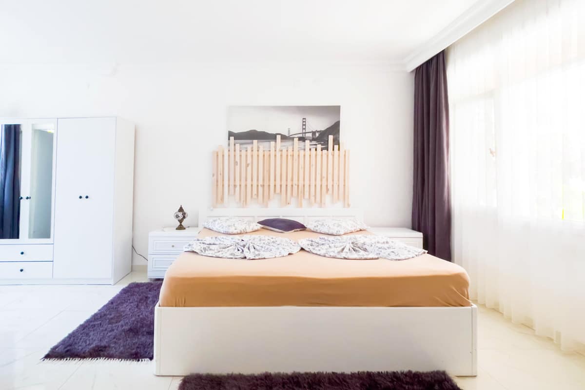 bedroom Bright and cozy bedroom featuring a large bed with a wooden headboard, soft neutral bedding, white furniture, and a large window with sheer curtains letting in natural light.
