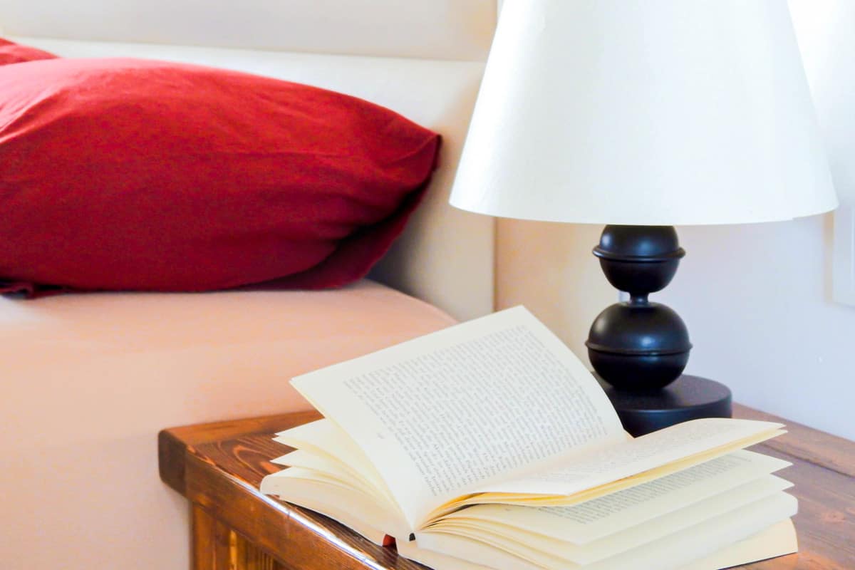closeup-book-room Cozy bedroom detail with an open book on a wooden bedside table, next to a black lamp with a white shade, and a bed with deep red pillows in the background.