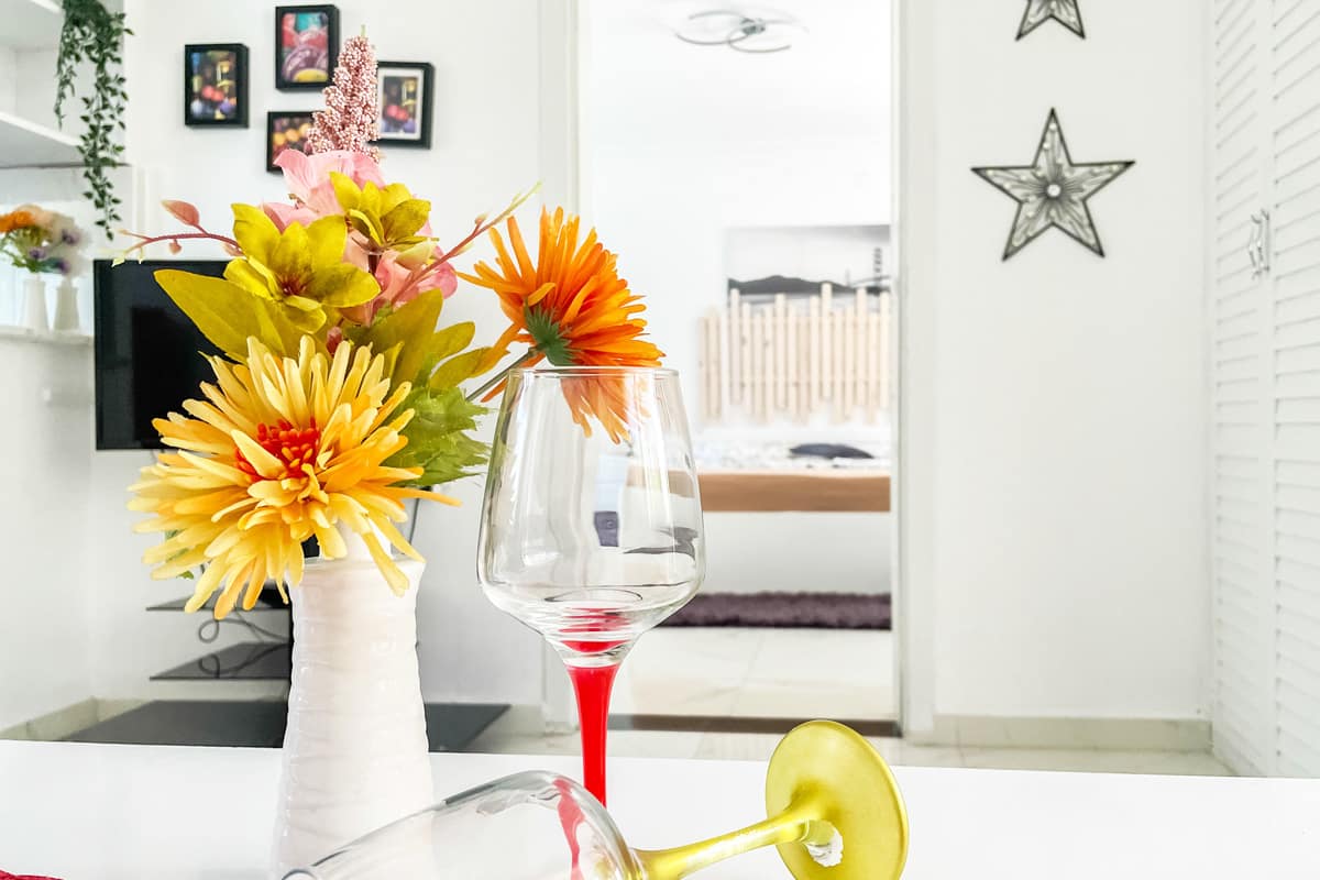 decor-room Elegant interior detail with a white vase of vibrant yellow and orange flowers, two decorative wine glasses, and a cozy bedroom visible through an open doorway in a bright, minimalistic setting.
