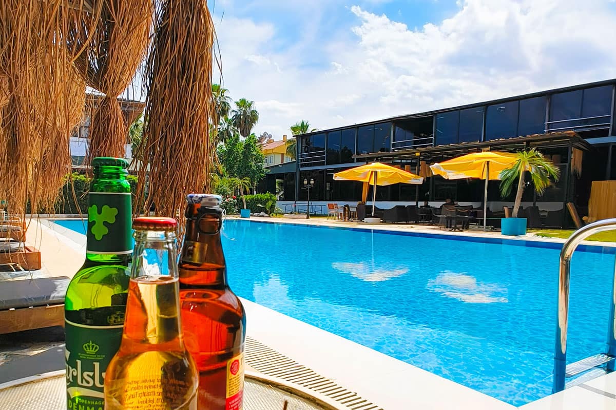 drinks-by-the-pool Refreshing poolside view with three bottles of beer on a table, overlooking a large blue swimming pool lined with sun loungers and bright yellow umbrellas. A modern glass building and palm trees complete the sunny resort setting.