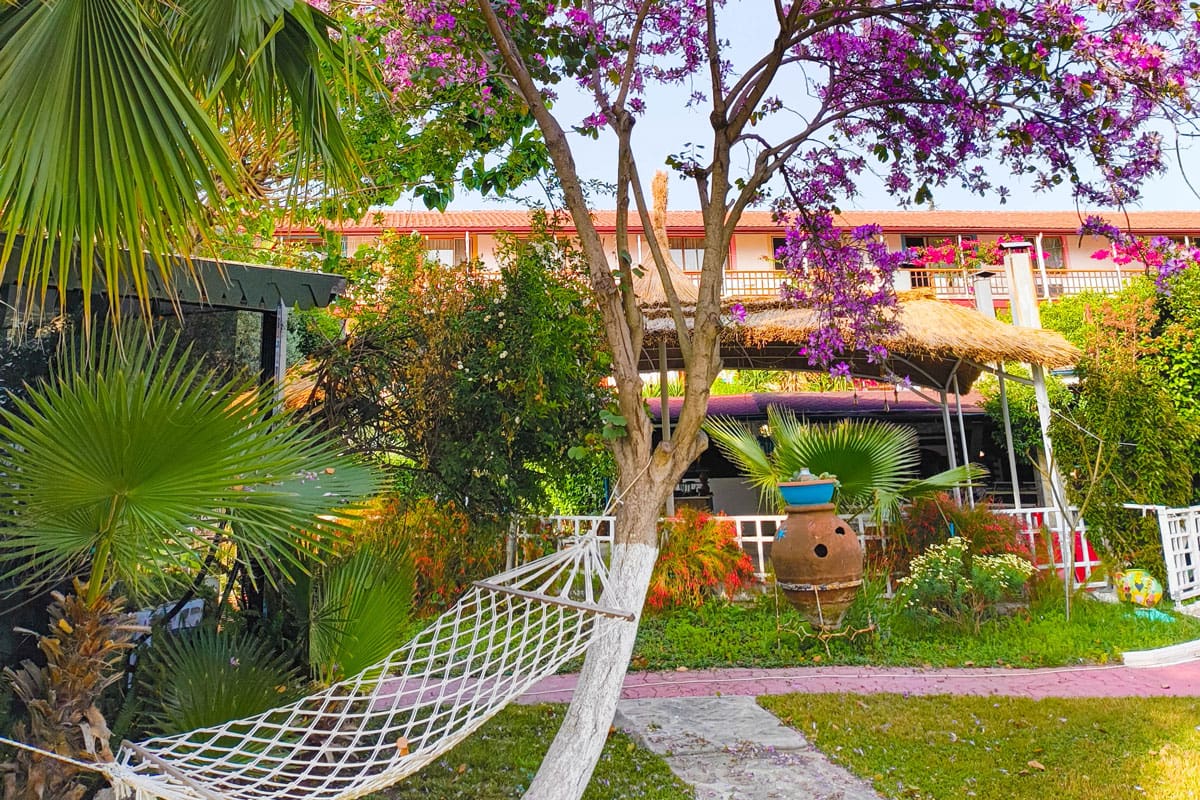 hammock Cozy hammock tied between trees in a lush garden filled with palm leaves, vibrant flowers, and a large terracotta pot. Behind the garden is a traditional building with red rooftops and a shaded terrace, creating a peaceful Mediterranean retreat atmosphere.