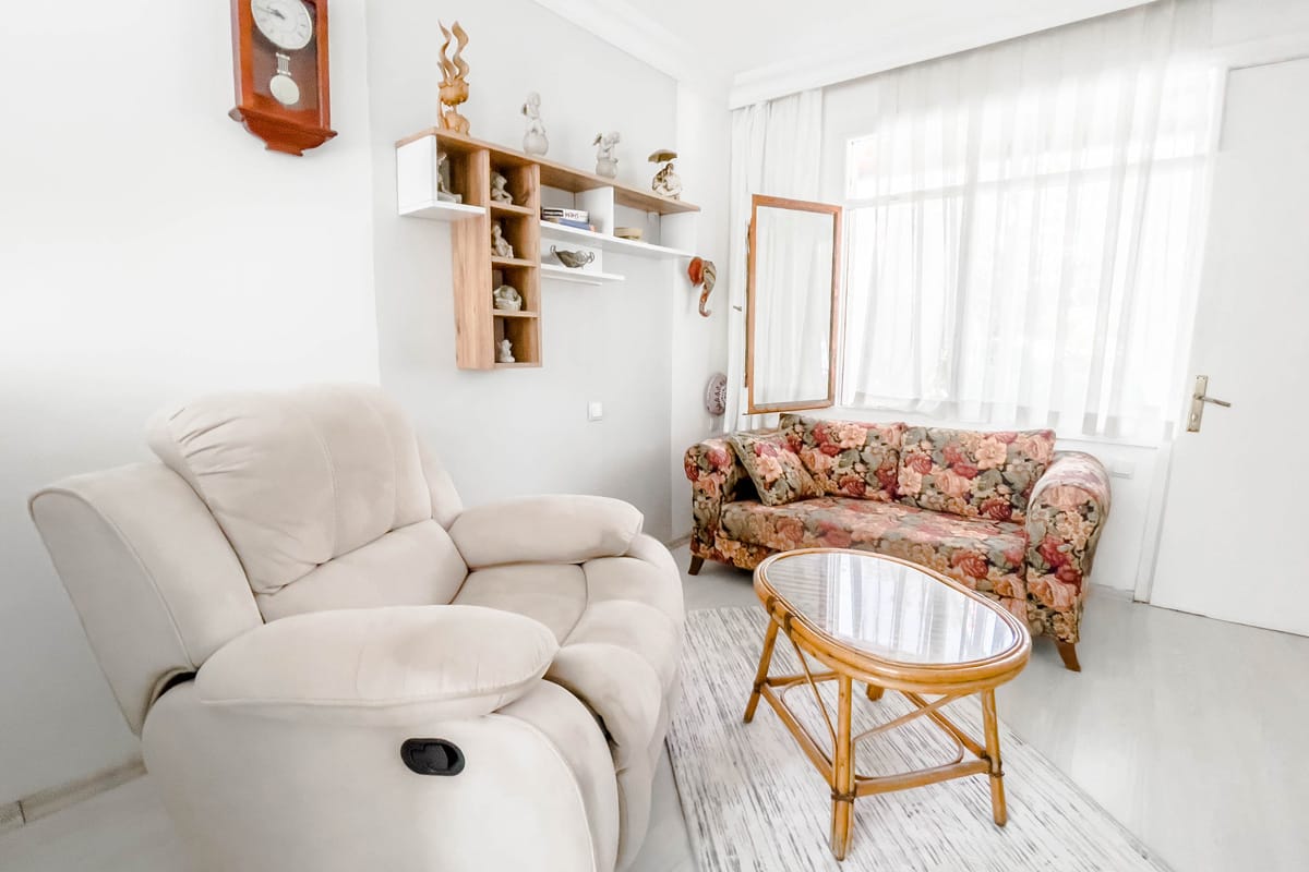 interior Cozy living room with a plush beige recliner, a floral-patterned sofa, a glass-top rattan coffee table, and wall-mounted shelves decorated with figurines, all softly lit by natural light through sheer curtains.