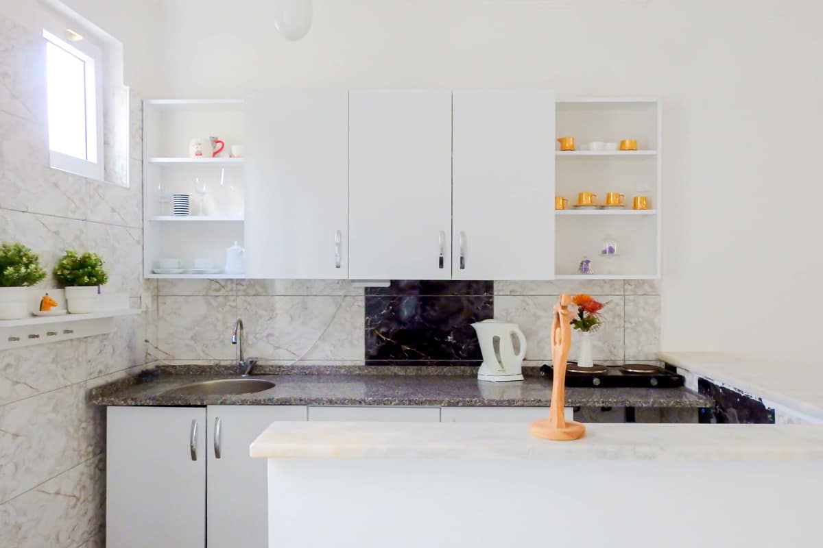 kitchenette Bright kitchenette with white cabinets, marble-patterned backsplash, open shelves displaying cups and dishes, and a countertop with a kettle and decorative vase.