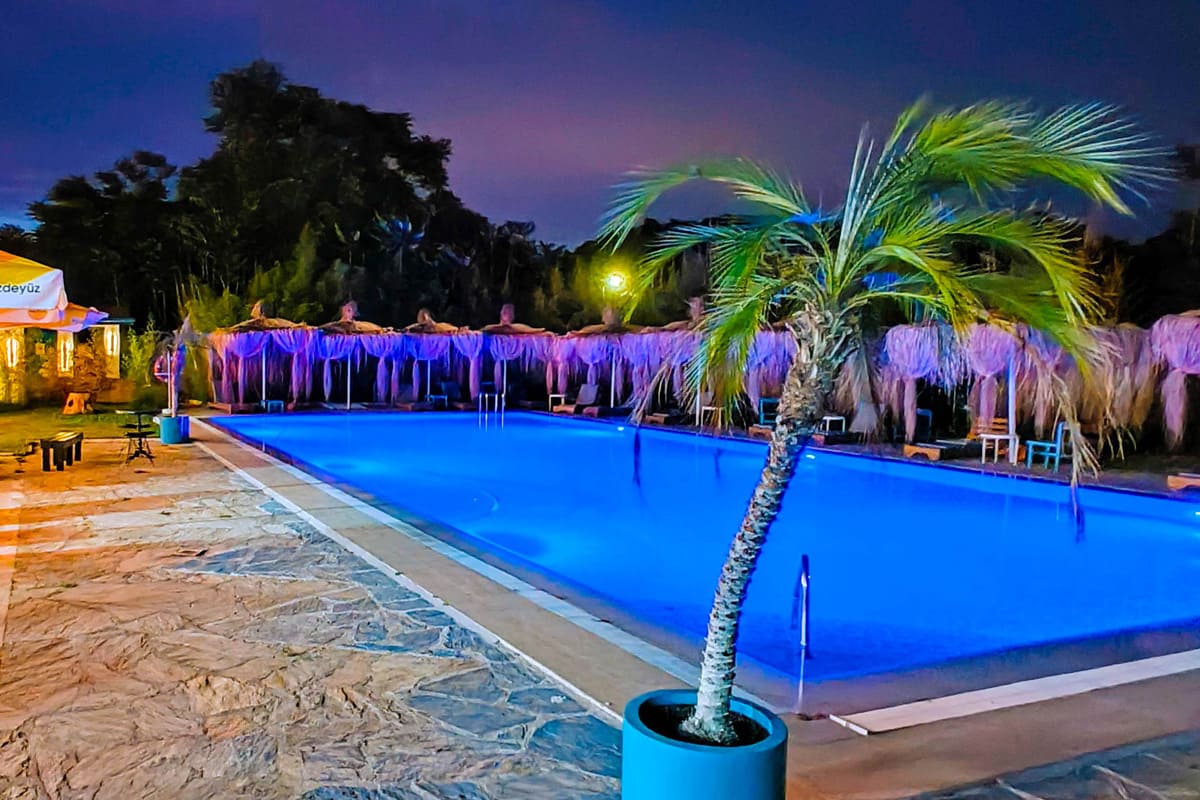 nighttime-pool Tranquil nighttime view of a glowing blue pool surrounded by thatched cabanas and a single potted palm tree, with warm lights casting a cozy ambiance against the dark sky.