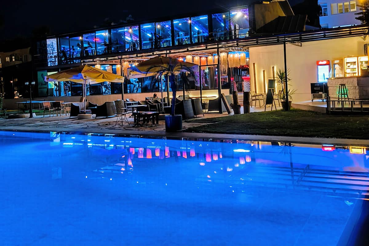 nighttime-poolside Night view of a glowing blue pool reflecting colorful lights from a modern two-story building with glass walls, cozy outdoor seating, and bright yellow umbrellas, creating a vibrant evening atmosphere.