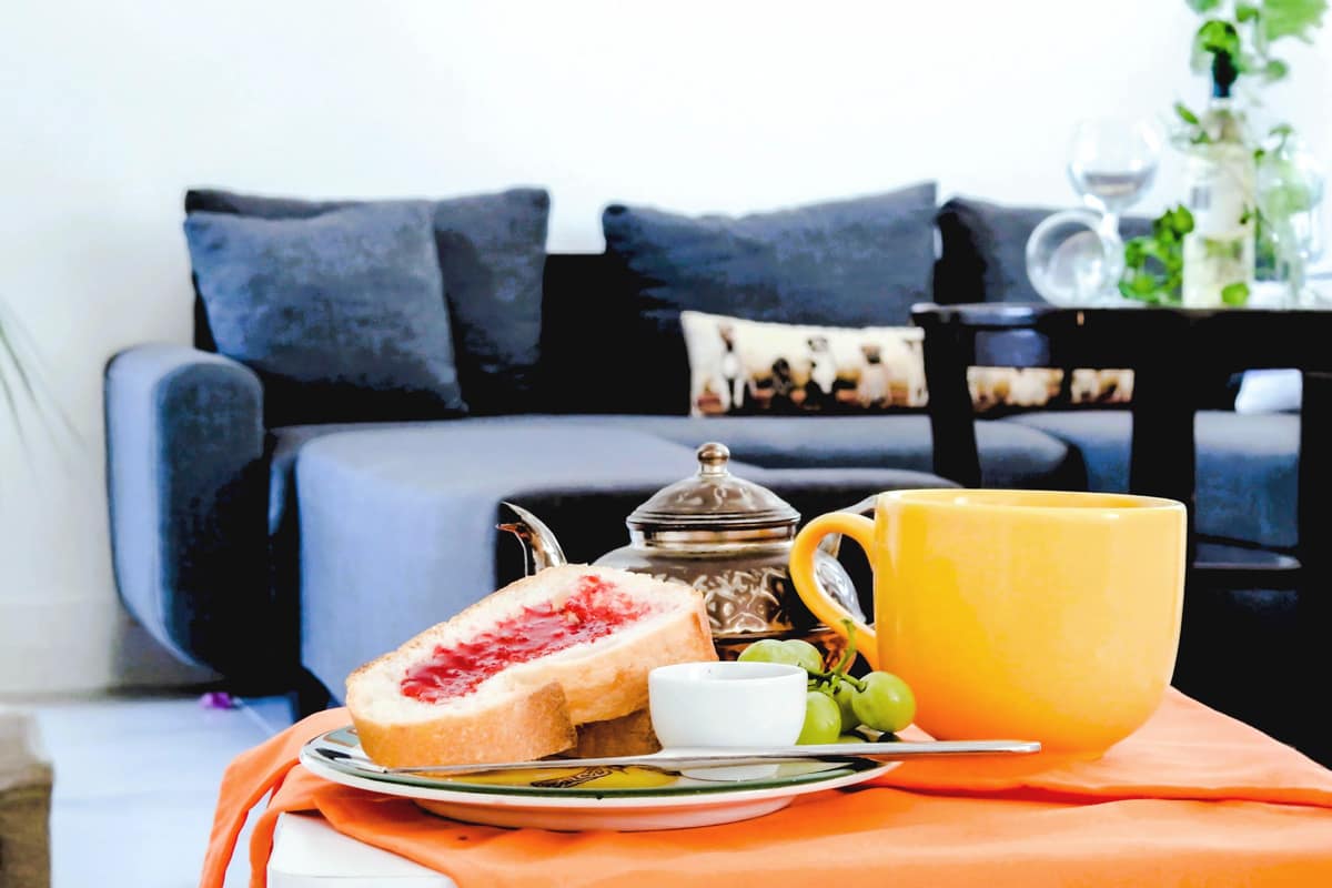 photoshoot-room Cozy breakfast setup with toast and jam, green grapes, a yellow mug, and a decorative teapot on a white table with an orange cloth, set in front of a modern gray sofa with patterned cushions.