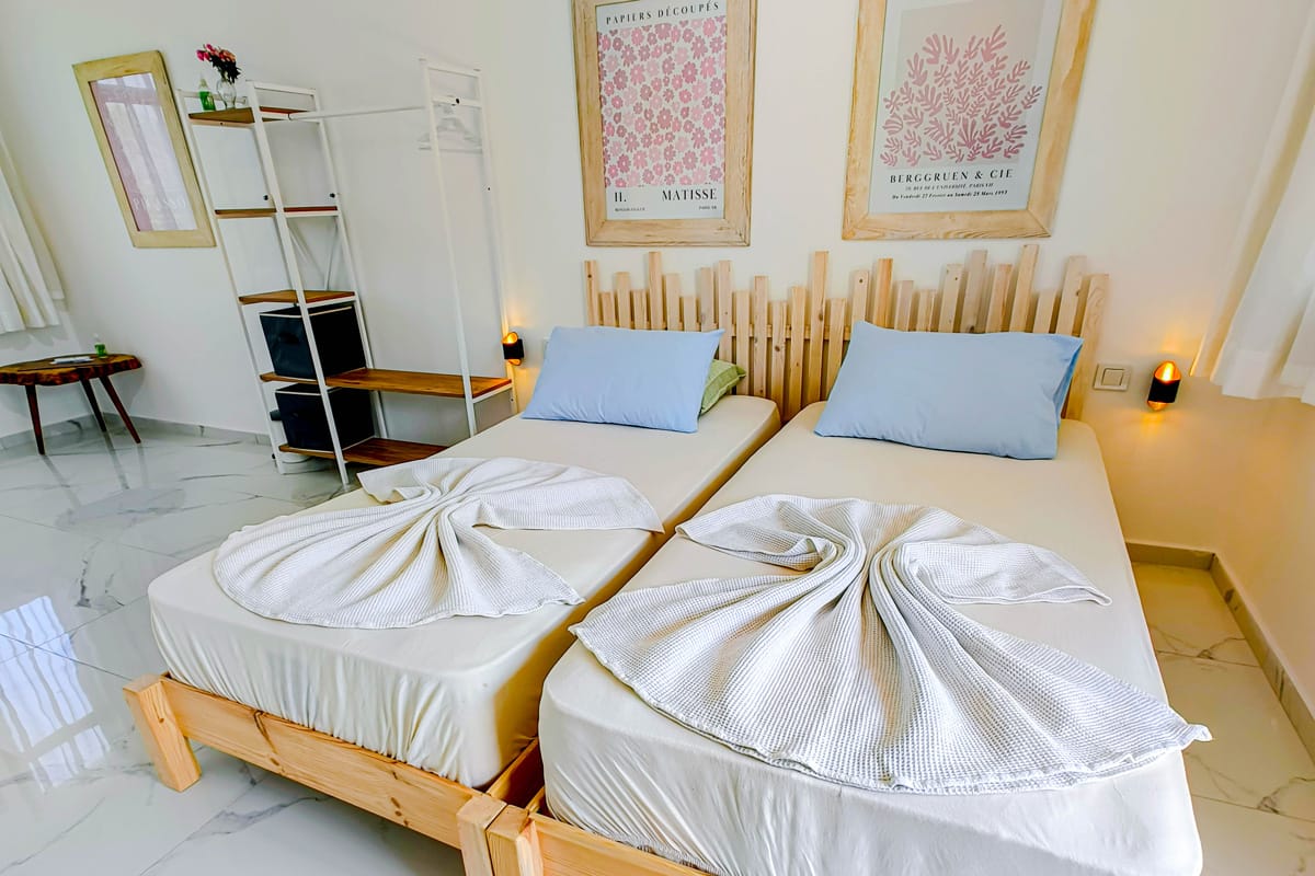 rooms-header Modern guest room with two twin beds featuring light wood frames, blue pillows, and towels folded into decorative shapes, complemented by framed wall art and minimalist furniture.